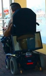 A wheelchair driving along with a computer monitor mounted on the back of the wheelchair showing a video of a bum in jeans 