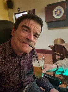 A head and shoulders shot of DrDazz sitting in his wheelchair in the hotel with someone someone's hand holding a glass of beer with a straw in it to him mouth. He looks satisfied.